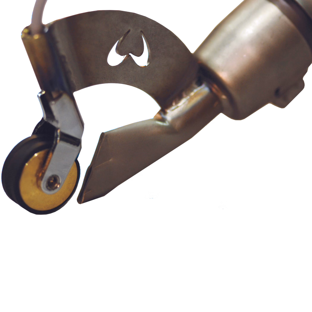 WOLFF_Dummy_47049_PUR welding nozzle_2025-08 Nahaufnahme eines metallischen Handwerkzeug-Elements: eine Stahlaufnahme mit einem kleinen Rollenrad und einer spitz zulaufenden, meißelartigen Düse. Das Bauteil zeigt Schweißnähte, eine gebogene Halterung mit einer herzförmigen Ausstanzung als Logo und gebürstete Metalloberflächen mit leichten Verfärbungen durch Hitze. Das Foto wurde vor weißem Hintergrund aufgenommen und fokussiert das Rad, die Halterung und die abgeschrägte Spitze; es deutet darauf hin, dass das Teil zum Führen, Trimmen oder Abdichten in der Produktion oder Textilverarbeitung verwendet wird.
