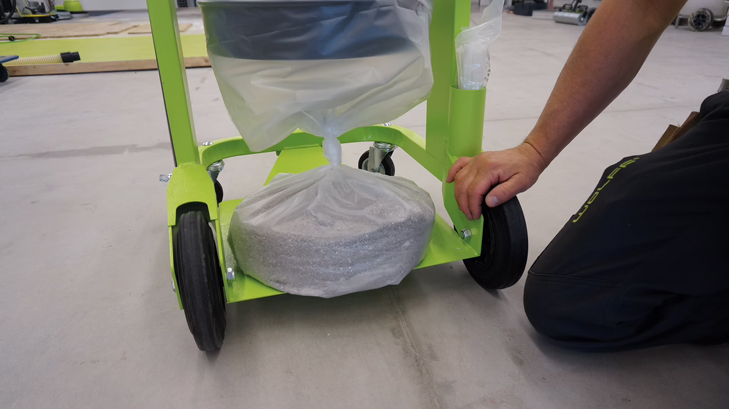 WOLFF_Application_Image_VacucleanLP_3 Eine Nahaufnahme eines grünen Wagens mit einer Plastiktüte, die am Boden mit Material gefüllt ist. Der Wagen hat zwei schwarze Räder, die auf dem Boden sichtbar sind, und die Hand einer Person ist zu sehen, die den Wagen oder seinen Inhalt anpasst. Im Hintergrund ist ein Arbeitsbereich mit verstreuten Werkzeugen und Materialien zu sehen, die auf ein laufendes Projekt hinweisen.