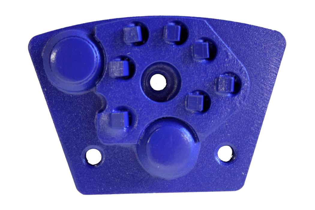 Nahaufnahme eines kleinen blauen Kunststoff- oder Metallbauteils vor weißem Hintergrund. Die trapezförmige Platte verfügt über zwei auffällige, runde Erhöhungen, ein zentrales rundes Loch, mehrere kleine quadratische Vorsprünge im oberen Bereich und zwei kleinere Befestigungsbohrungen an der unteren Kante. Die Oberfläche ist leicht strukturiert und die Farbe ist ein kräftiges Kobaltblau.