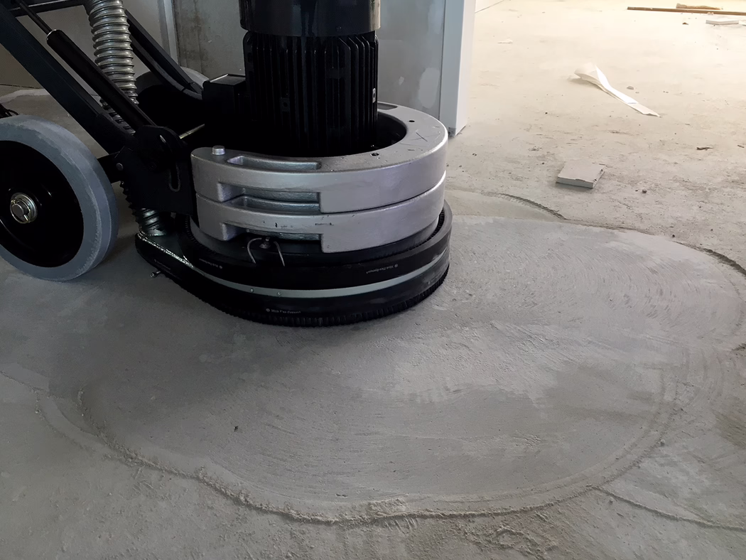 WOLFF_Application_Image_Neo400_1 Nahaufnahme einer Bodenschleifmaschine, die eine Betonoberfläche auf einer Baustelle glättet; sichtbar sind das Motorgehäuse, der runde Schleifkopf und die Räder sowie Schleifspuren und Staub auf dem grauen Betonboden bei kühlem Innenlicht. Keine Personen sichtbar.