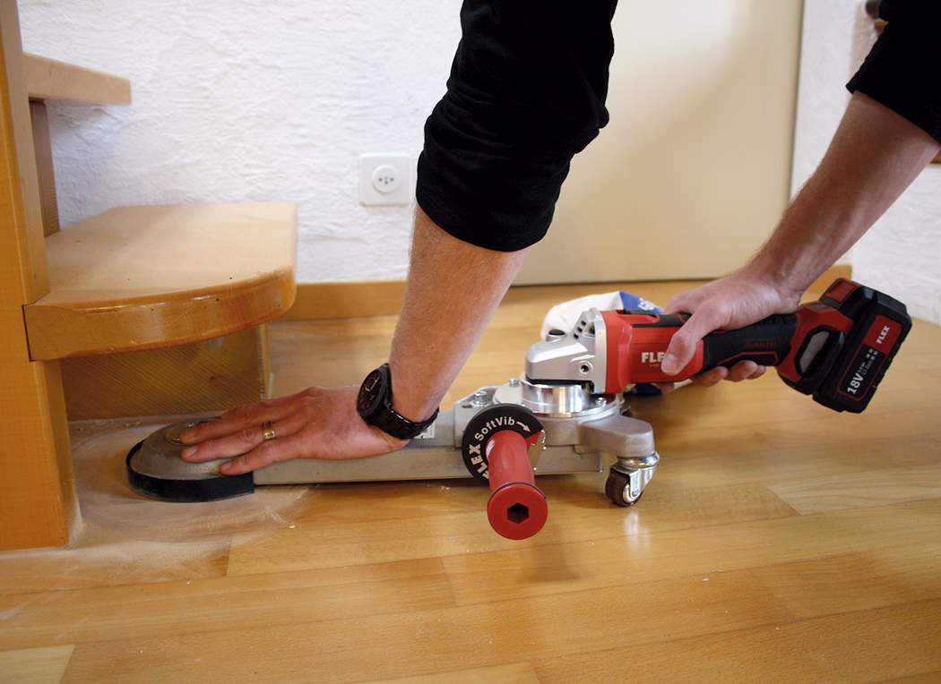 Derendinger_Application_Image_URM-DS_Akku_2 Nahaufnahme einer Person, die neben einer Treppenstufe einen Holzboden abschleift. Ein rotes, akkubetriebenes FLEX-Winkelschleifergerät ist mit einer Flachanschlussplatte und einer Schleifscheibe montiert; die rechte Hand hält den Griff, die linke Hand stabilisiert die Platte am Boden. Unter dem Metalladapter ist eine Lenkrolle sichtbar, feiner Holzstaub liegt auf dem Parkett, und an der Wand sind eine Steckdose sowie eine Holzfußleiste zu sehen. Auf dem linken Handgelenk ist eine Armbanduhr, am Finger ein Ehering; die Szene vermittelt eine sorgfältige Innenrenovierung am Stufenrand.