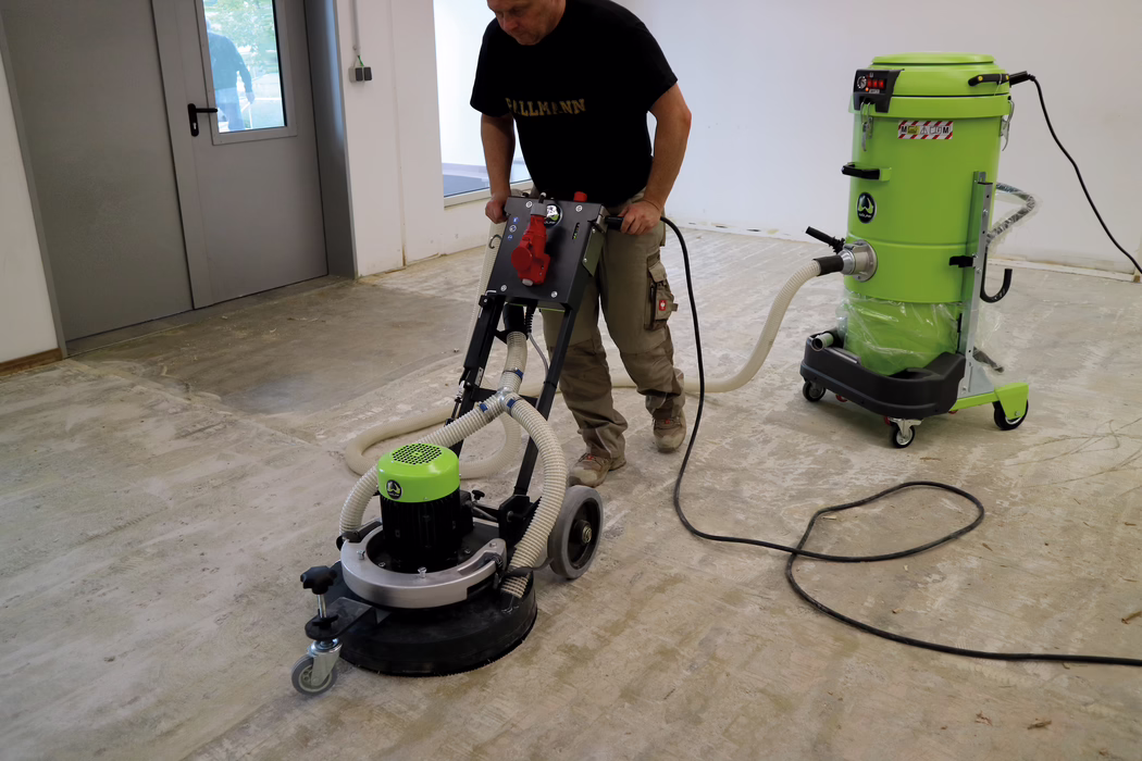 WOLFF_Application_Image_Neo400_3 Ein Handwerker bedient eine Bodenschleifmaschine, die an einen hellgrünen Industriesauger angeschlossen ist; Schläuche und Stromkabel liegen auf dem nackten Betonboden eines leeren Raums, der für Renovierungsarbeiten vorbereitet wird.