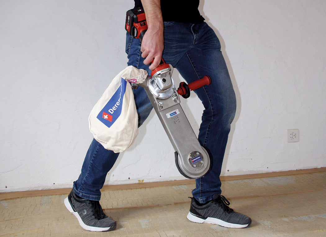 Derendinger_Application_Image_URM-DS_Akku_3 Eine Person, nur von der Taille abwärts sichtbar, trägt blaue Jeans und schwarze Turnschuhe und hält einen handgeführten Bodenschleifer mit Staubsack. Das Gerät hat einen roten Griff und ein Metallgehäuse; am Stoff-Staubsack ist ein blaues Logo mit weißem Kreuz zu sehen. Im Bild sind ein Holzfußboden und eine weiße Wand zu sehen.