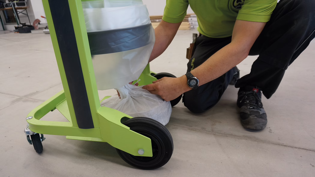 WOLFF_Application_Image_VacucleanLP_2 Eine Person, die neben einem grünen Wagen mit Rollen kniet. Der Wagen hat einen Müllsack in einem Halter, und die Person bindet den Müllsack zu. Die Umgebung ist eine Werkstatt mit sichtbaren Werkzeugen im Hintergrund.