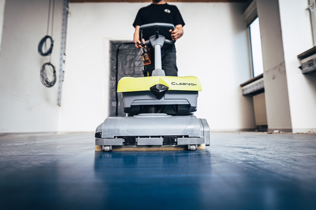 WOLFF_Application Image_Clean Fox_2021-10 Nahaufnahme einer gelb-grauen Bodenreinigungsmaschine mit der Aufschrift "Cleanfox", die von einer Person in einem Innenraum während Renovationsarbeiten bedient wird; niedrige Perspektive, blauer Boden und unscharfer Hintergrund mit Kabeln.